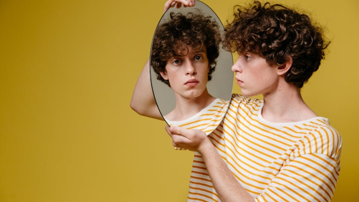 A young man with curly hair holds a mirror reflecting his serious expression against a yellow backdrop.